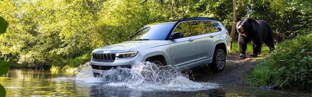 2026 Jeep Grand Cherokee riding in a water bear is behind and surrounded by forest.