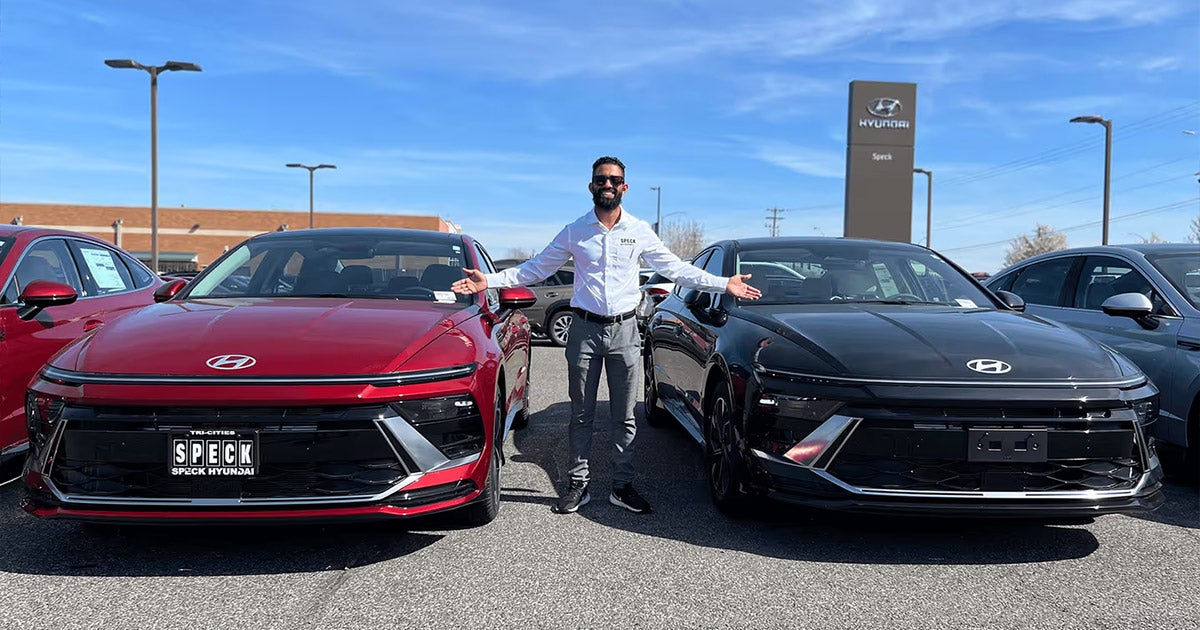 Two Hyundai sedans, one red and one black, parked at a Hyundai dealership with a salesperson standing between them