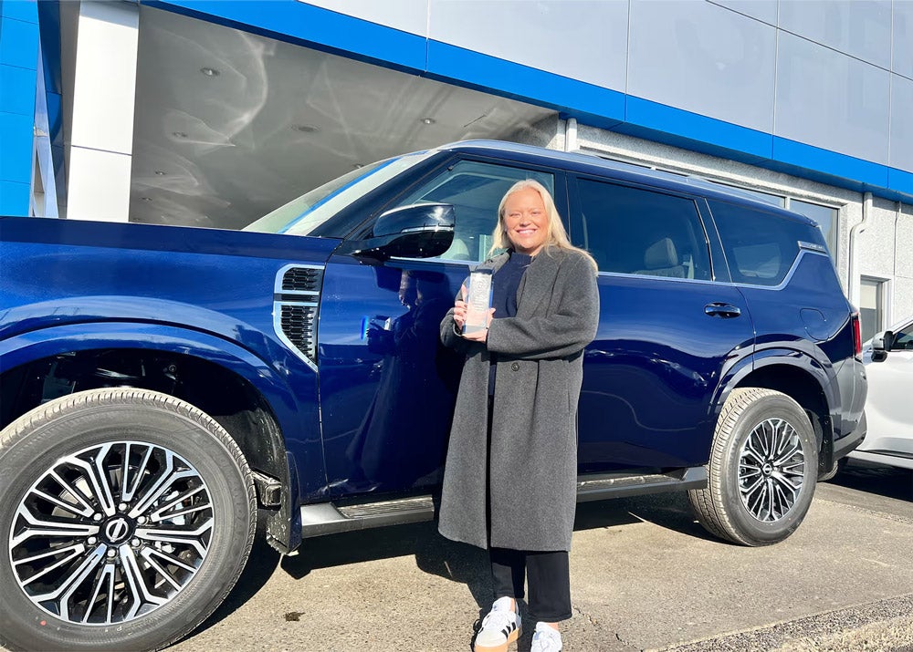 Blonde woman in gray coat standing next to a blue SUV, holding a white cup at a car dealership