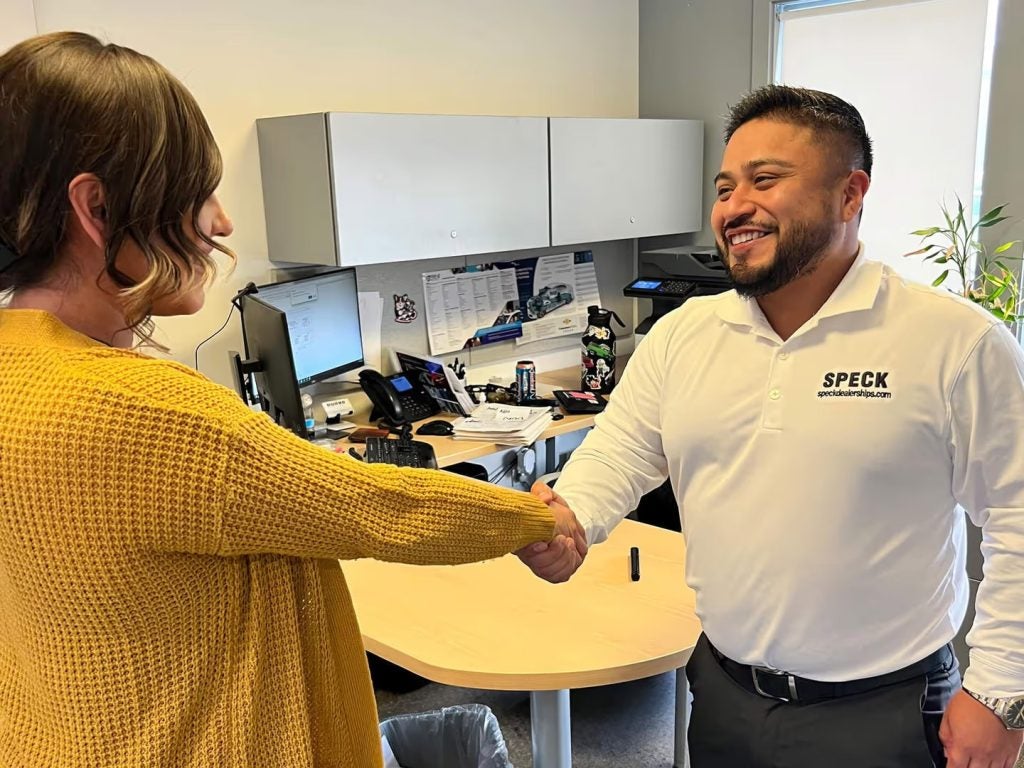 Business professionals shaking hands in an office, one wearing a yellow sweater, the other in a white Speck polo shirt.
