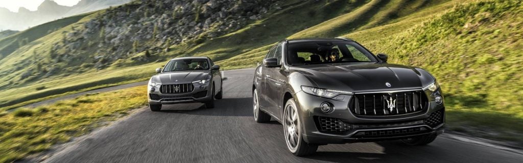 Two Maserati SUVs driving on a winding mountain road, with scenic green hills and rocky terrain in the background.