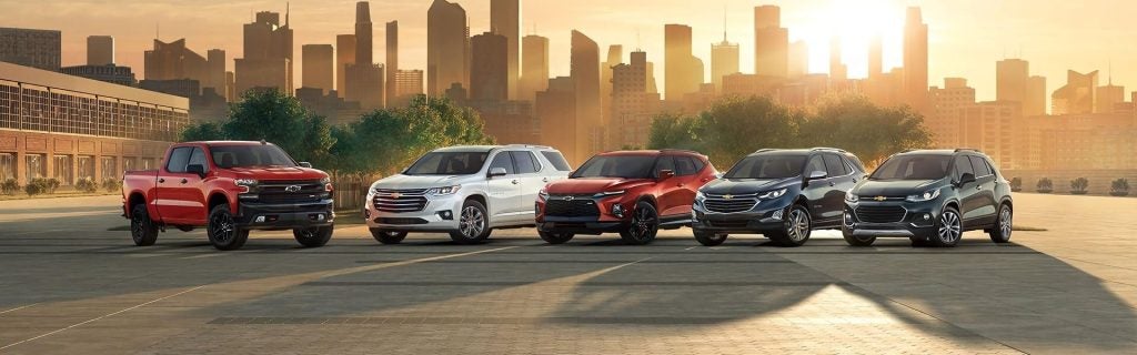 A lineup of five diverse Chevrolet vehicles parked in a row, with a golden city skyline at sunset in the background.
