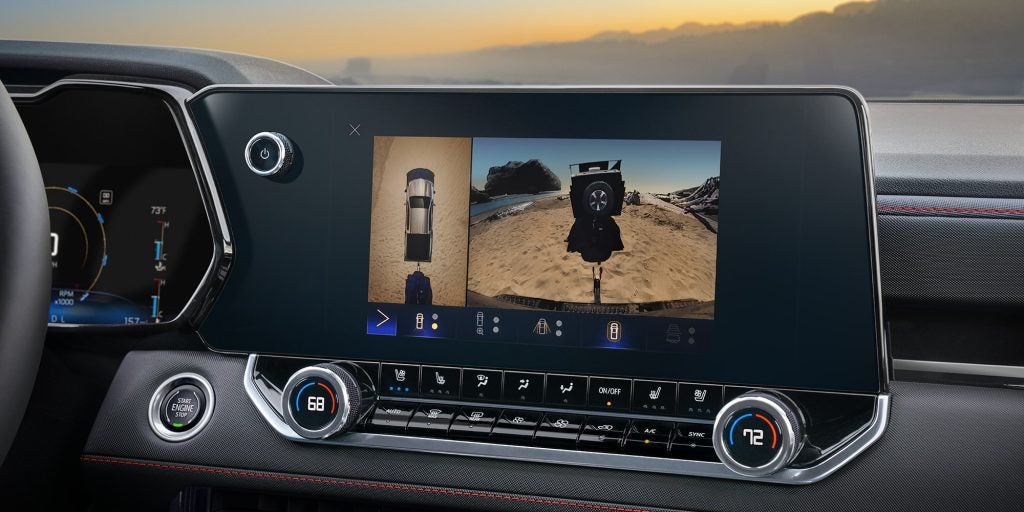 The interior of a modern vehicle featuring a digital touchscreen displaying split camera views of a truck towing a trailer on sandy terrain, surrounded by advanced climate controls and a textured dashboard with red stitching.