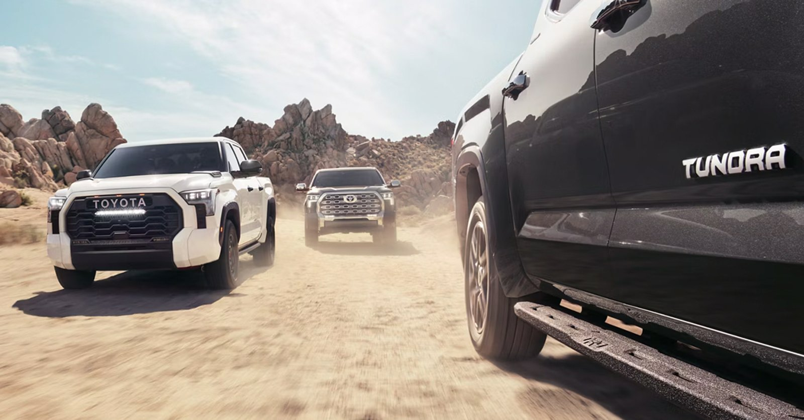 A white and a gray pickup truck on a desert plain, with two motocross riders in the foreground.