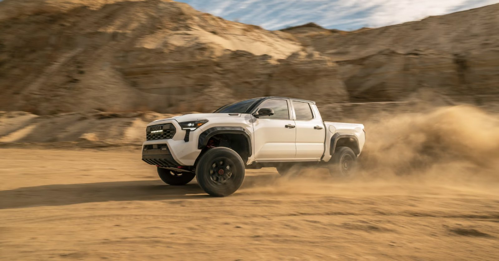 Rear view of a white Toyota Tacoma pickup truck with a bed rack, driving on a rocky dirt path.