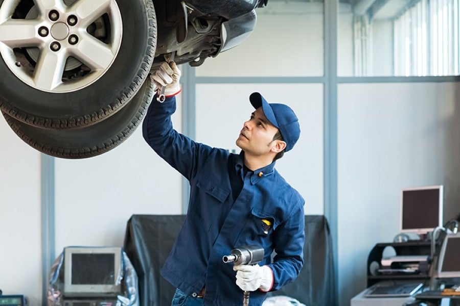 Technician inspecting the underside of a raised vehicle, checking the suspension or wheel area while holding a power tool in an auto repair shop.