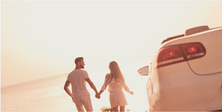 Couple holding hands walking toward the beach at sunset beside a white SUV, with warm golden lighting and a coastal backdrop.