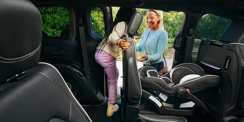 Interior view of a vehicle showing a child climbing into the back seat while an adult assists, with a rear-facing child car seat installed