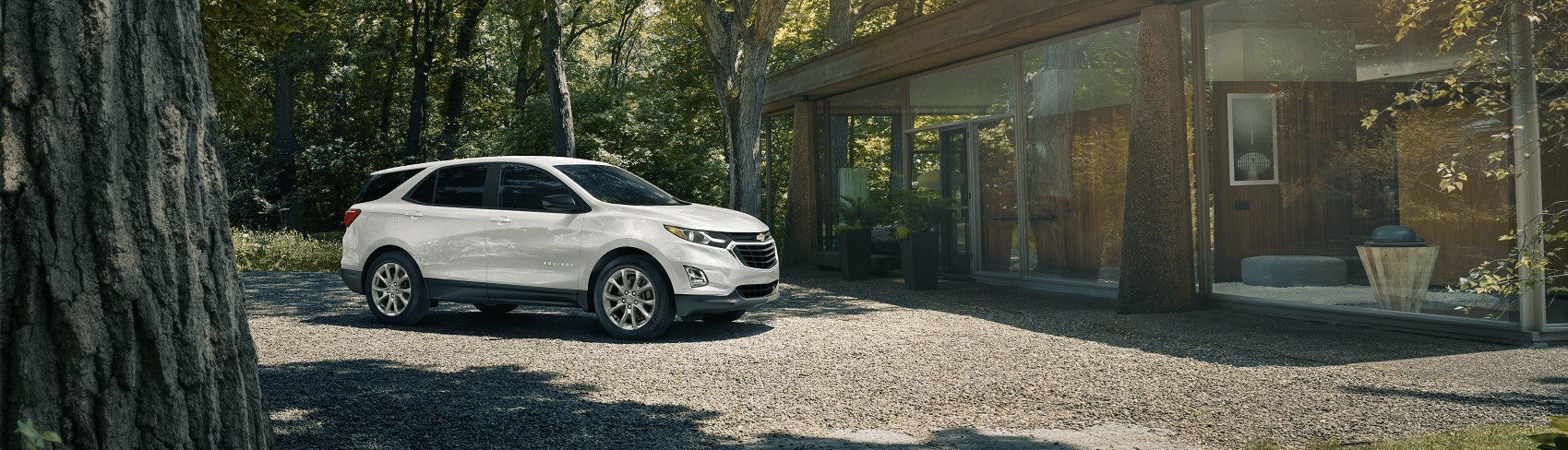 A shiny, new White Chevy Equinox is parked in sunshine outside of a stylish cabin in the woods