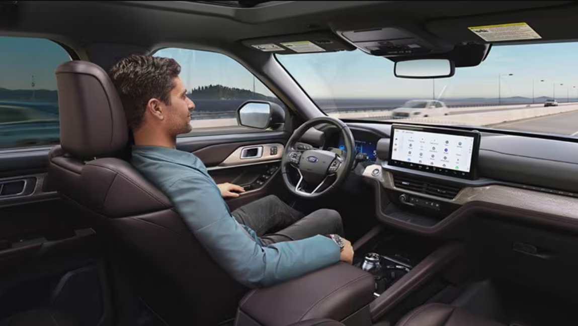 Driver seated behind the wheel using hands-free driving assistance on a highway, with a large touchscreen display visible inside a modern vehicle cabin.