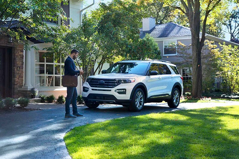 A man with a bag approaches a white Ford parked in a sunny driveway. The scene is tranquil, with green grass and trees surrounding a suburban house.