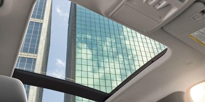 A modern car interior with a panoramic sunroof, offering a view of tall glass buildings above.