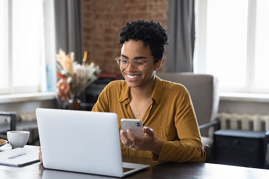 A person using a smartphone while working on a laptop at a desk in a home office setting.