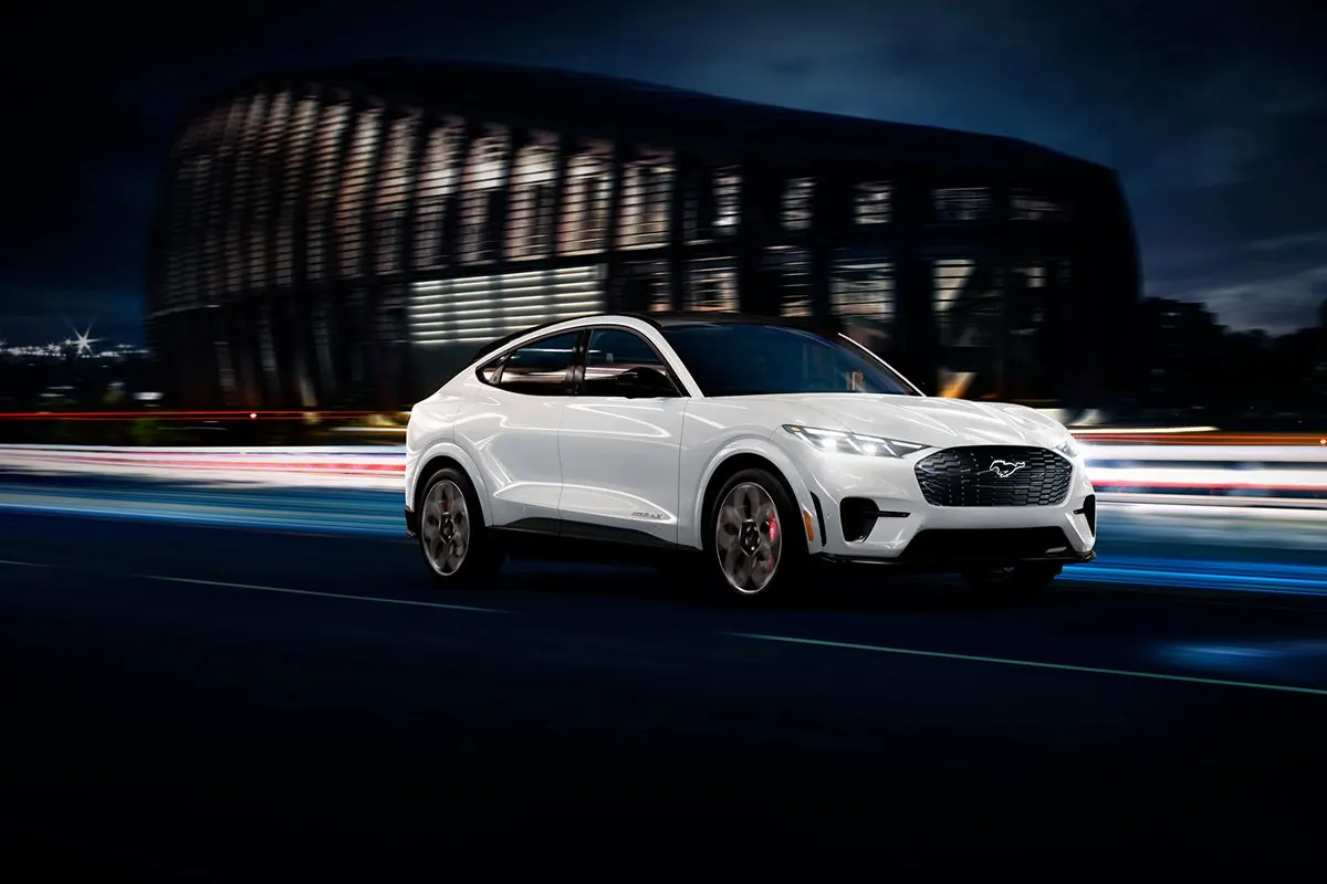 White Ford Mustang Mach-E driving past a lit arena at night