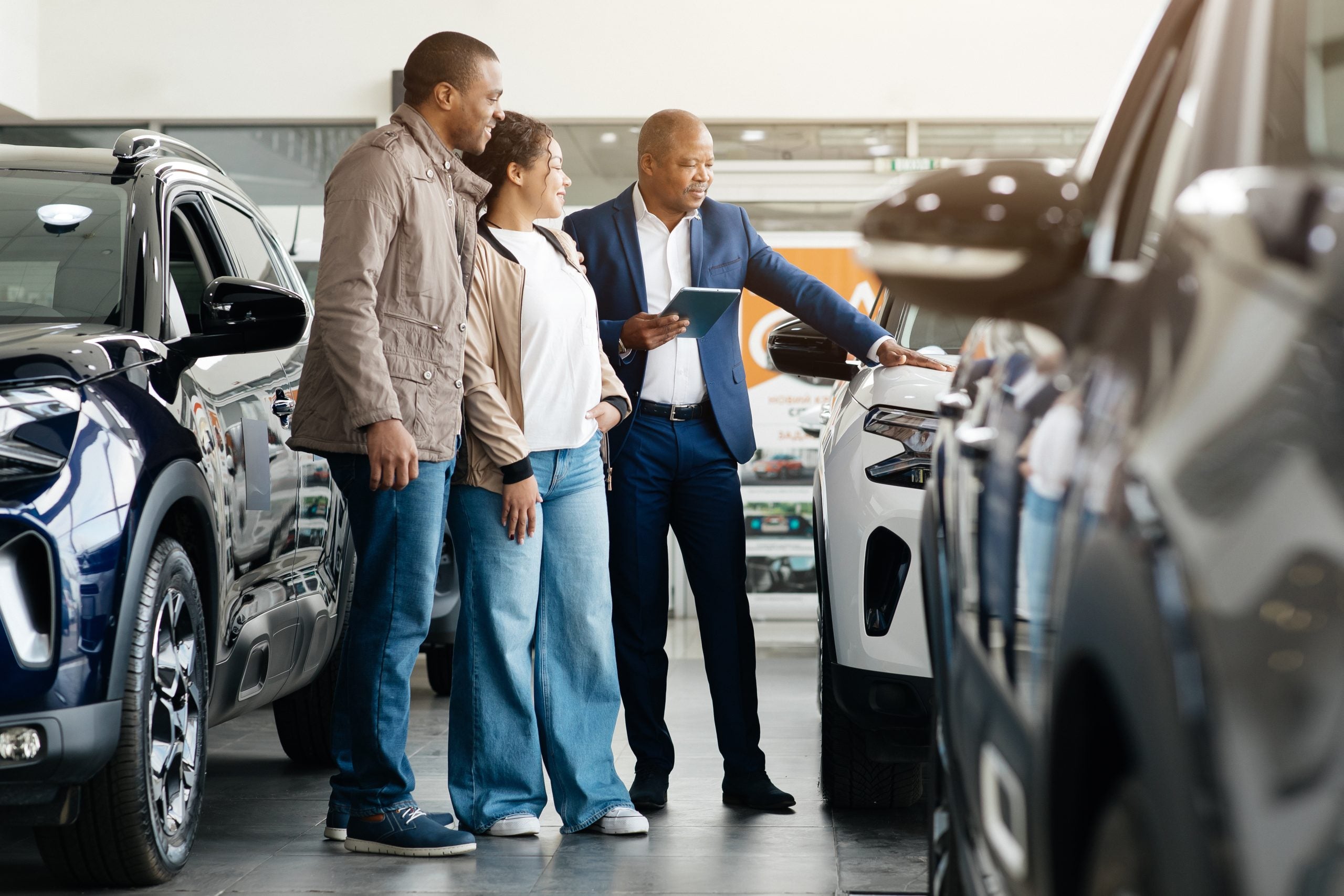 Car Selling Business. Manager Showing Luxury Automobile To Afro Spouses In Automobile Dealership Center. Panorama