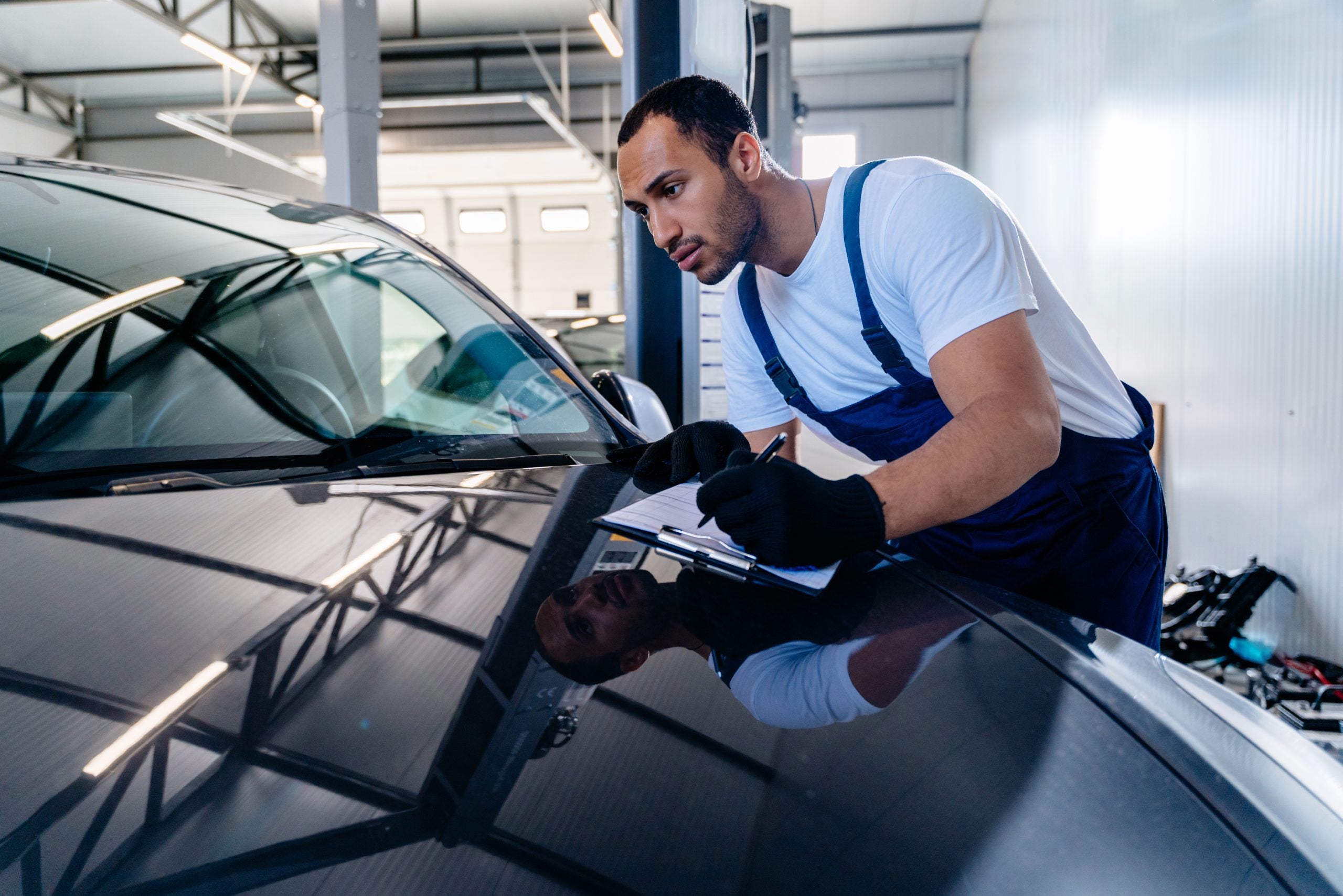 Arabian male auto mechanic or painter specialist professional car mechanic man in blue overalls white t-shirt with clipboard with papers document work in modern vehicle repair shop workshop indoor.