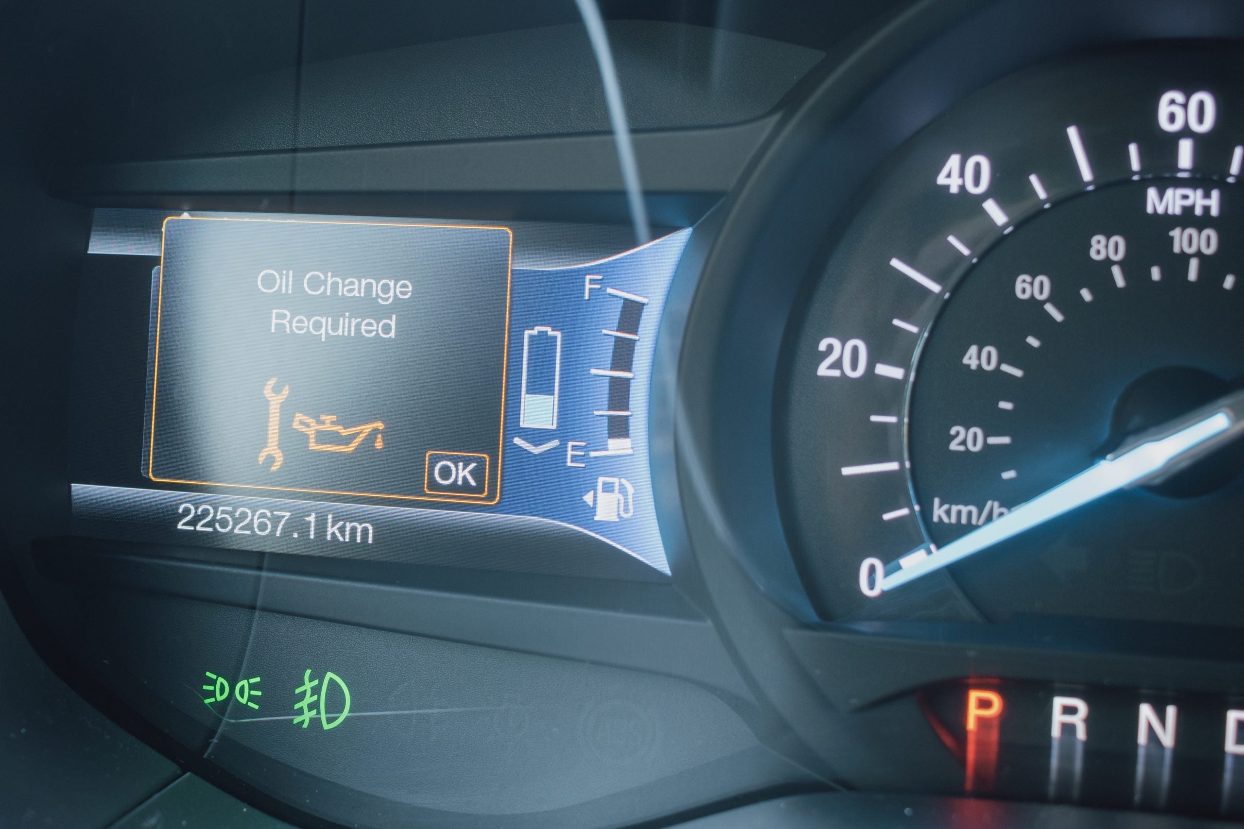 Close-up of a modern car dashboard, displaying an Oil Change Required warning light and message. Digital gauge cluster showing speedometer, fuel and battery status with high mileage
