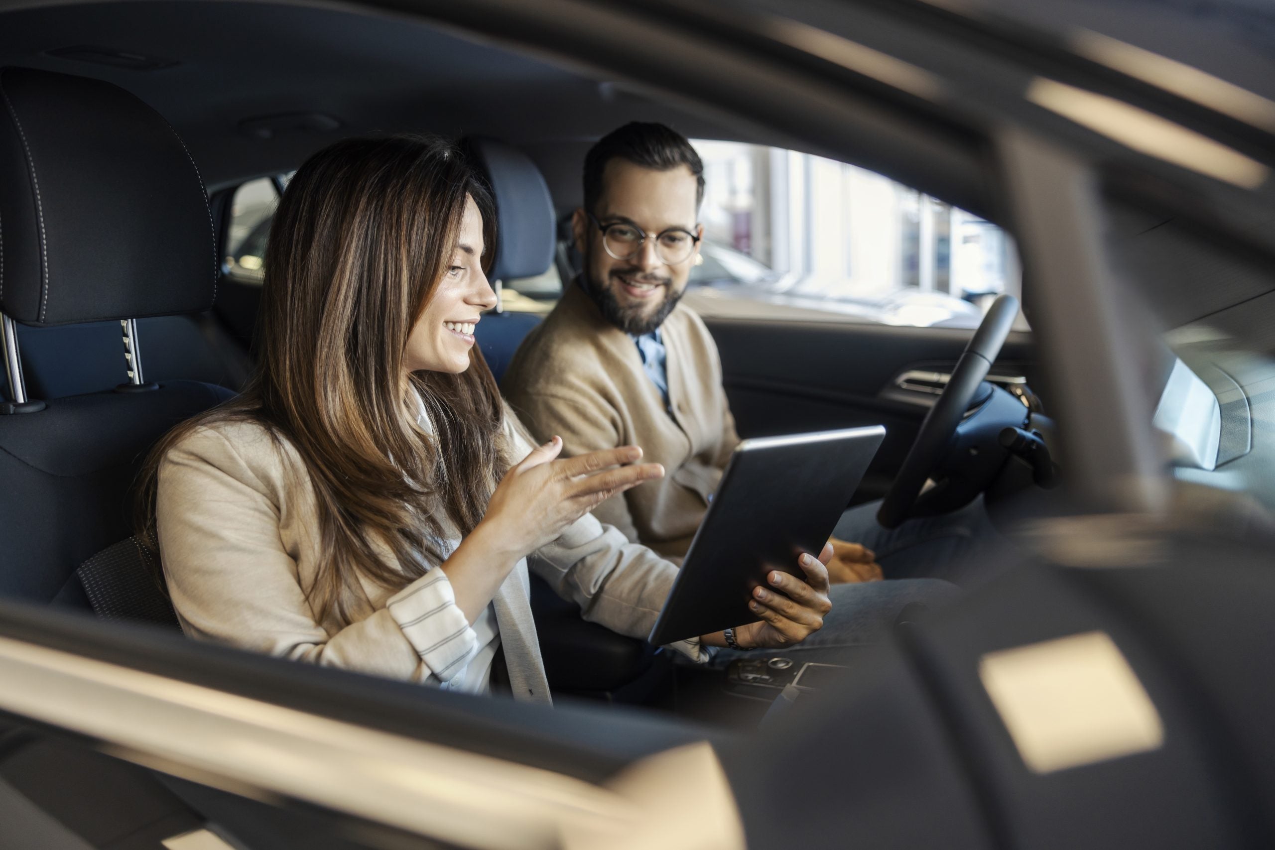 Buyer and saleswoman sitting in new car with tablet in hands and discussing price at car showroom.