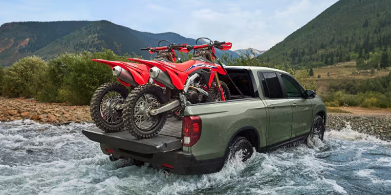 Green pickup truck driving through a shallow river carrying two red dirt bikes in its bed with mountains in the background.