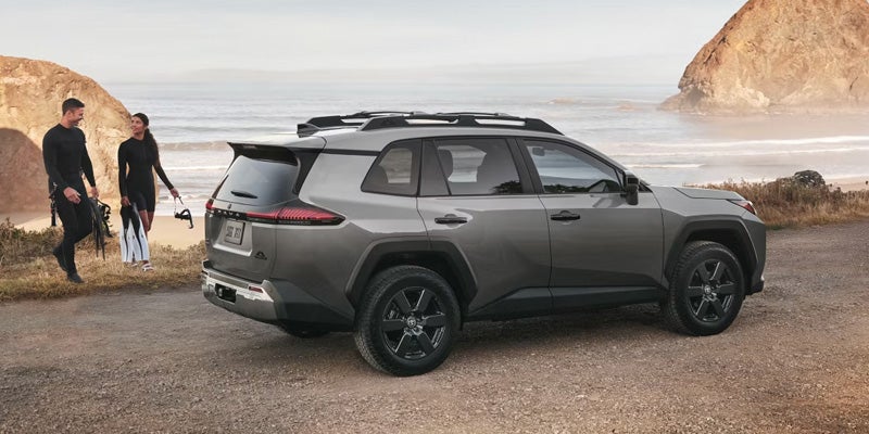 Gray SUV parked near beach with two people carrying surfboards