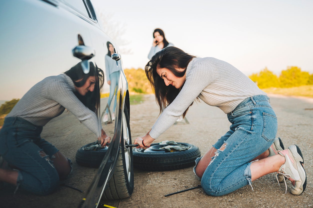 Woman changing a flat tire on the side of the road while her friend calls Savage L&B for a tow to their service center