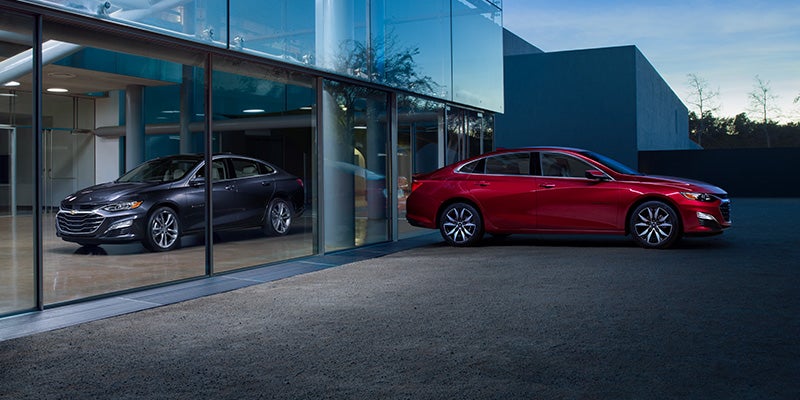 Two all new 2024 Chevrolet Malibu's, one black and one red, parked in front of a glass wall on a modern building. - Ted Britt Chevrolet Sterling, VA