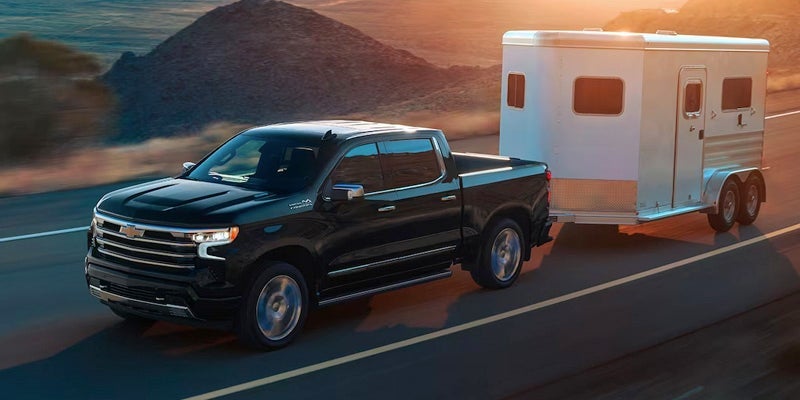 An all-new Black 2024 Chevy Silverado 1500 towing an animal trailer on the highway next to some rolling hills. - Ted Britt Chevrolet Sterling, VA