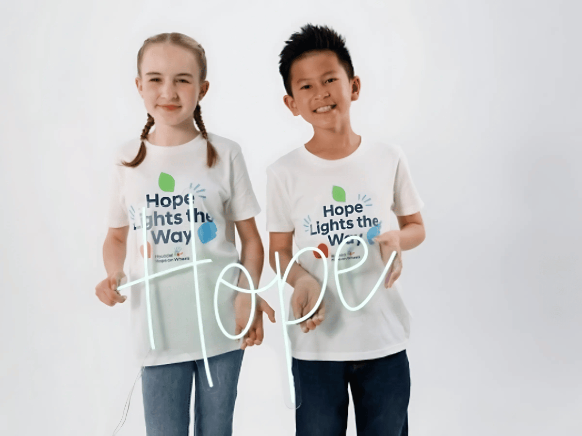 Two children wearing “Hope Lights the Way” Hyundai Hope On Wheels t-shirts, smiling and holding hands with the word “Hope” glowing in front of them.