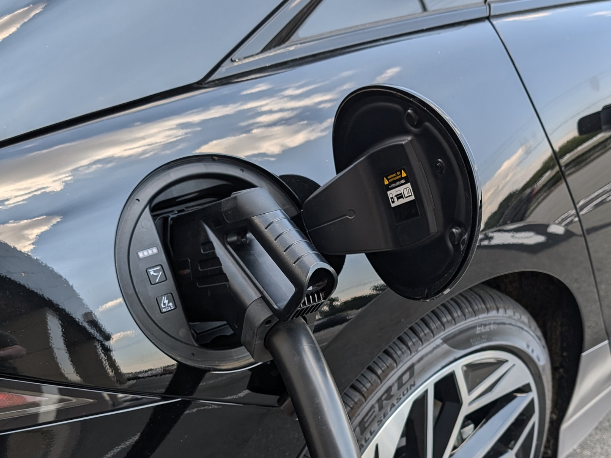 Close-up of a 2025 Hyundai Ioniq 5 charging at an EV station with a charging plug connected, reflecting the sky on its glossy black paint.