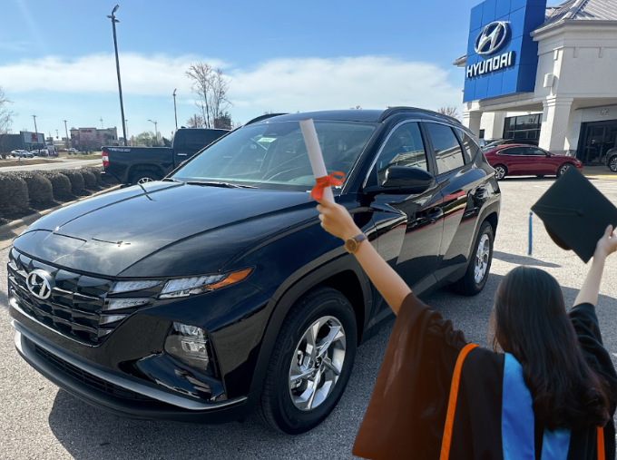 Excited graduate holding diploma and cap in front of a black Hyundai SUV at Crain Hyundai of Bentonville.