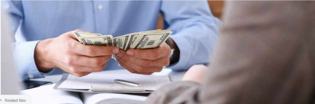 Hands holding a stack of U.S. dollar bills during a financial discussion at a desk with paperwork, suggesting a payment or transaction process.