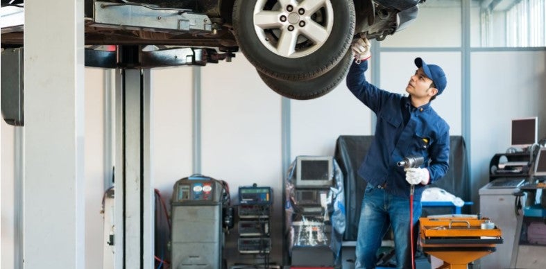 A mechanic working on a car lifted on a hydraulic lift in an auto repair shop, inspecting the undercarriage while using a tool.