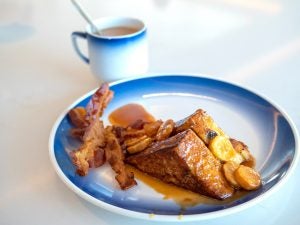 A plate of French toast, topped with caramelized banana syrup, with two strips of bacon. A cup of coffee.