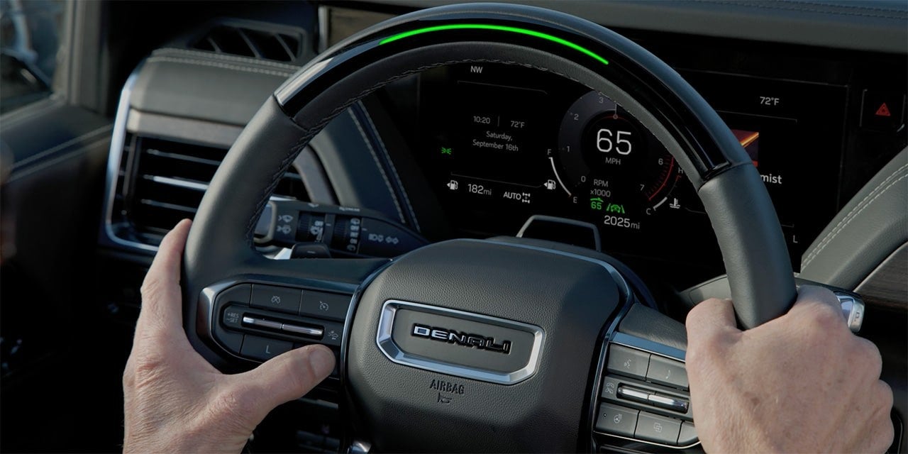 Hands on the steering wheel of a GMC Yukon with the green Super Cruise light bar lit