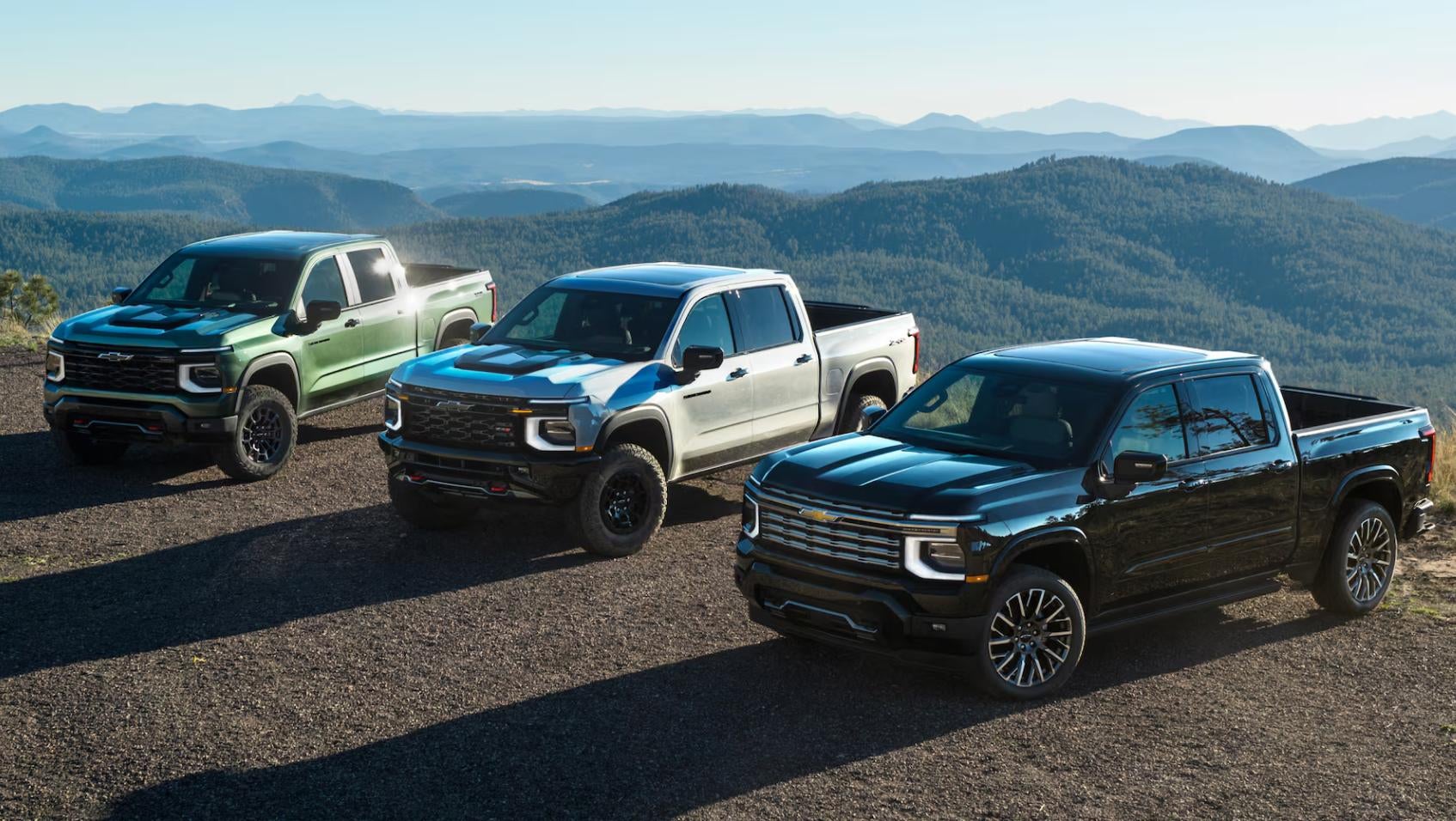 Three 2027 Chevy Silverado 1500 trucks in green, silver, and black parked on a mountain overlook