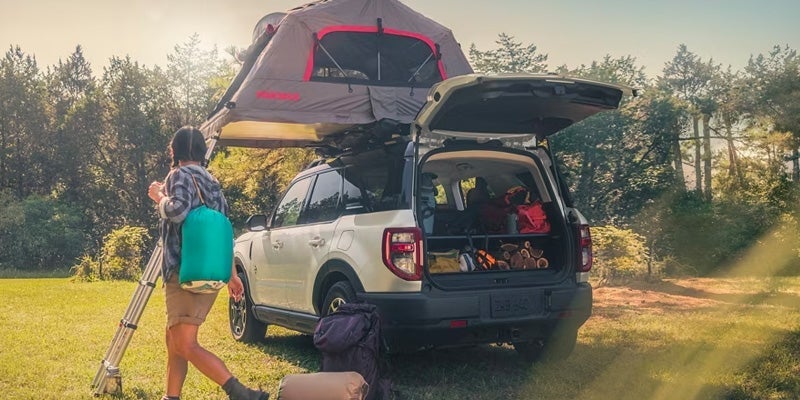 A person walking away from a white 2024 Ford Bronco Sport with the trunk open, exposing camping gear and a tent secured to the roof of the vehicle that is parked on grass on a sunny day