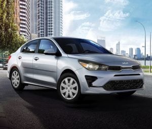 Profile view of a parked, silver 2022 Kia Rio with buildings in the background. | Kia dealer in Sherwood, AR.