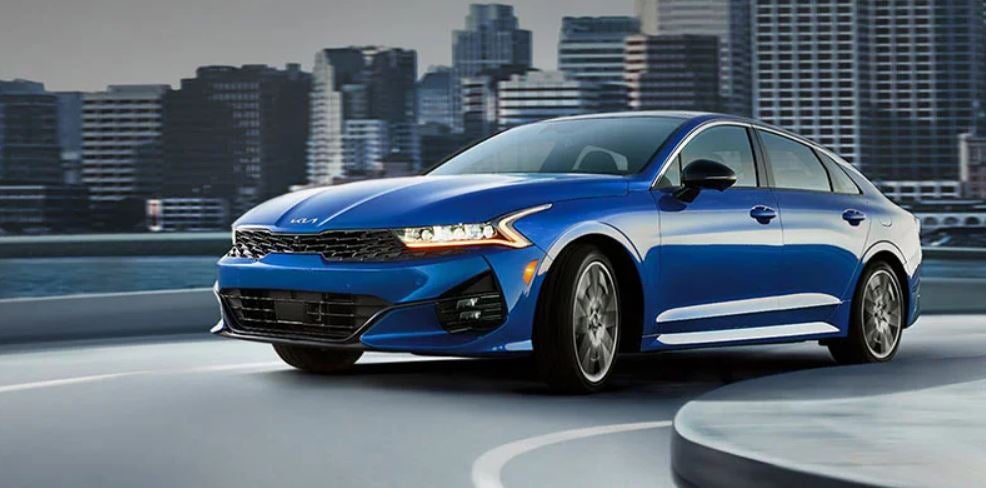 A blue 2022 Kia K5 being driven on the highway with tall buildings in the background. | Kia service in Conway, AR.