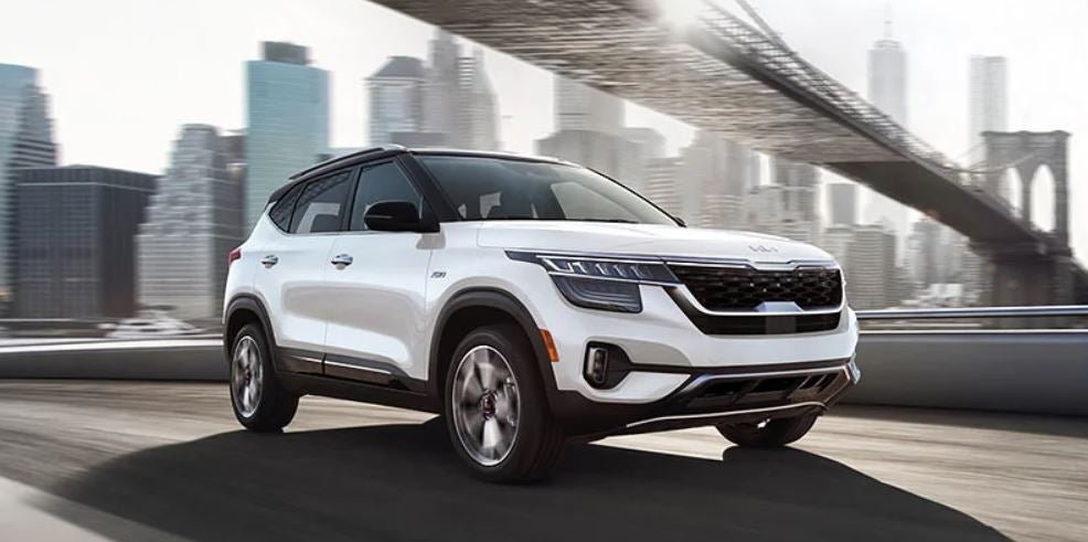 A white 2022 Kia Seltos being driven on the highway with a bridge overhead and buildings in the background. | Kia service center in Conway, AR.