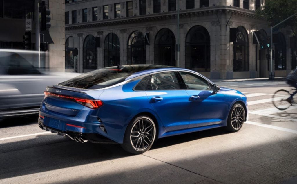 A blue 2022 Kia K5 being driven on the road with buildings in the background. | Kia dealer in Bentonville, AR.