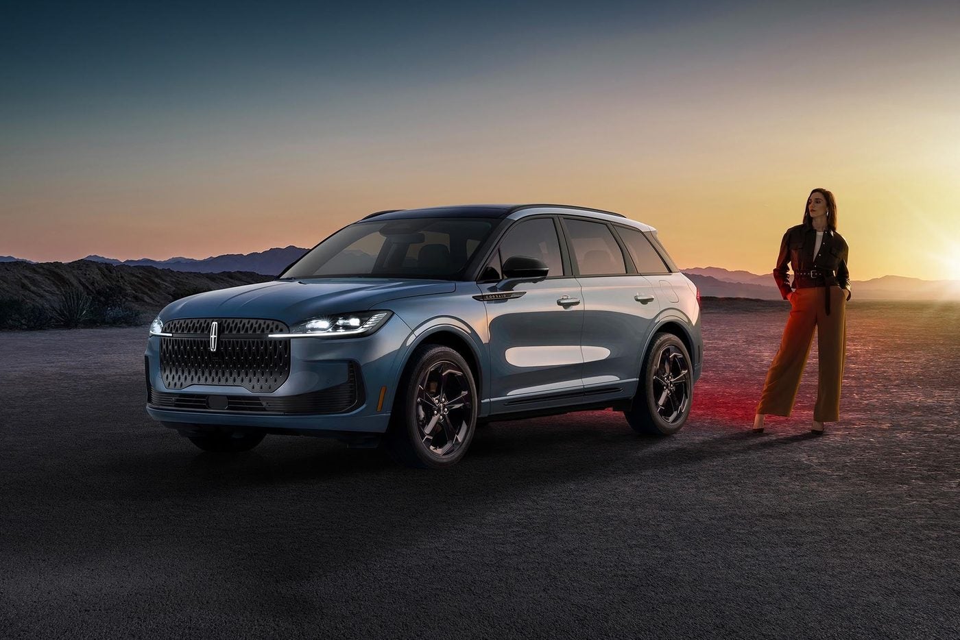 Blue gray Lincoln Corsair parked on a desert road at sunset with a woman standing beside it