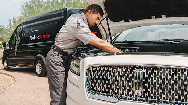 Lincoln technician in a gray uniform working under the hood of a white Lincoln next to a black Mobile Service van