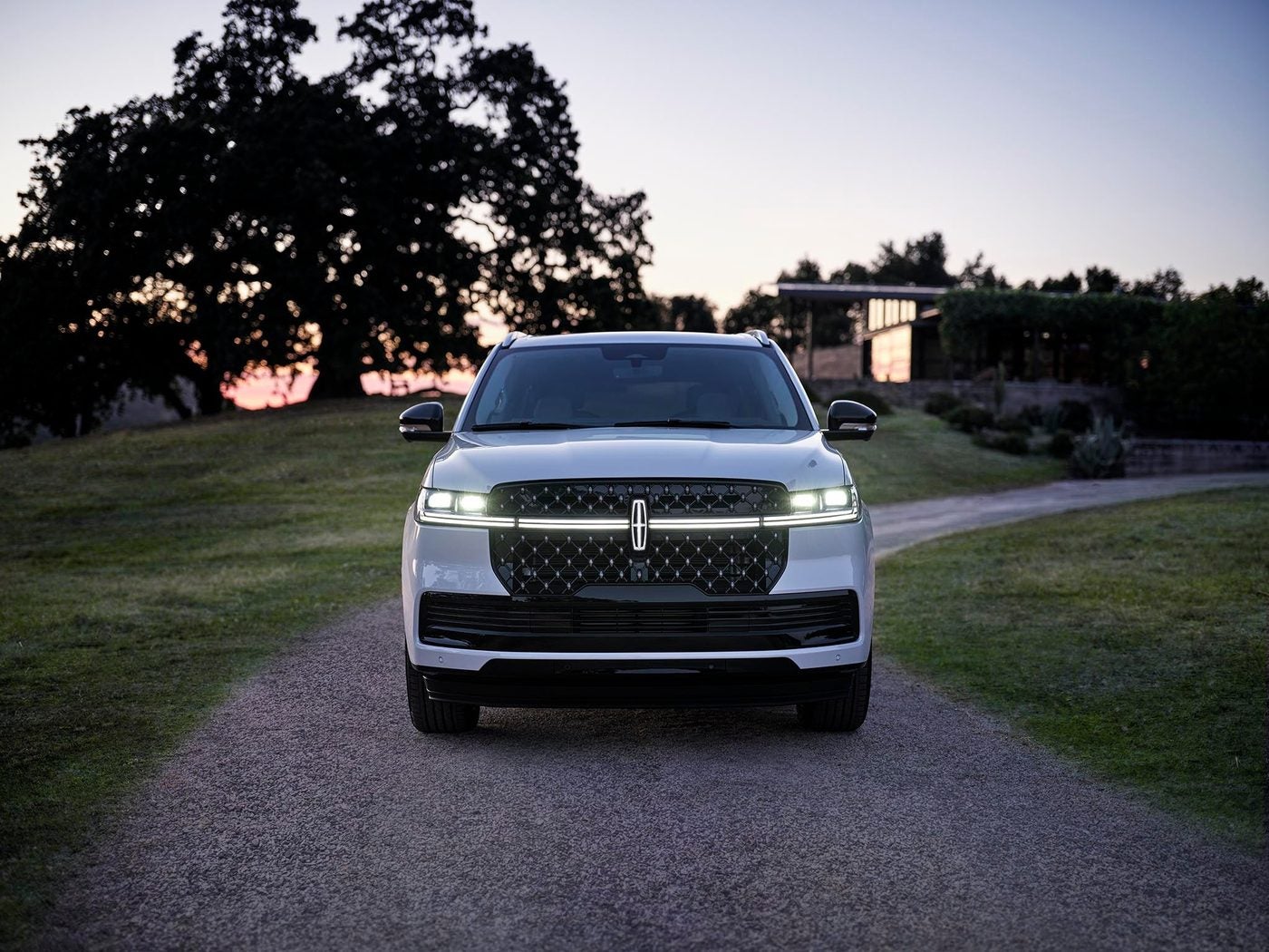 White Lincoln Navigator with headlights on, parked on a gravel driveway at dusk
