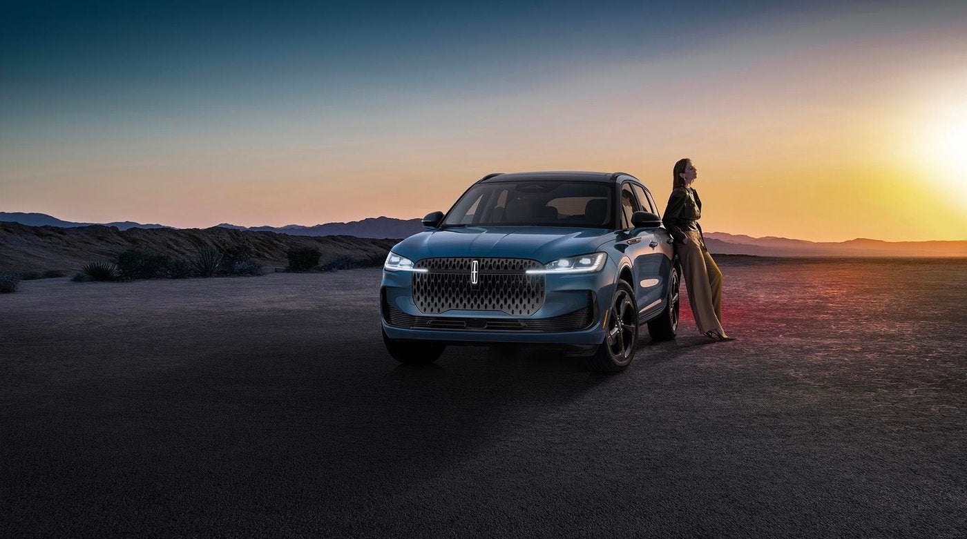 Woman leaning against a blue Lincoln Corsair parked outdoors at twilight