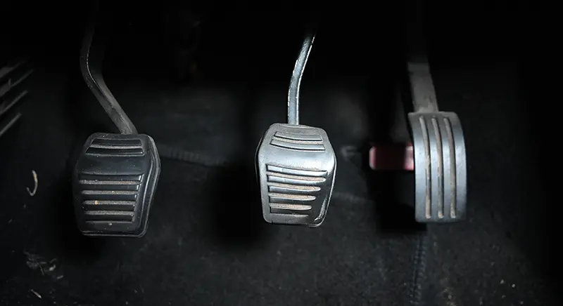 Three dirty, scratched, dark grey pedals on a dark car floor representing the clutch, brake, and accelerator.