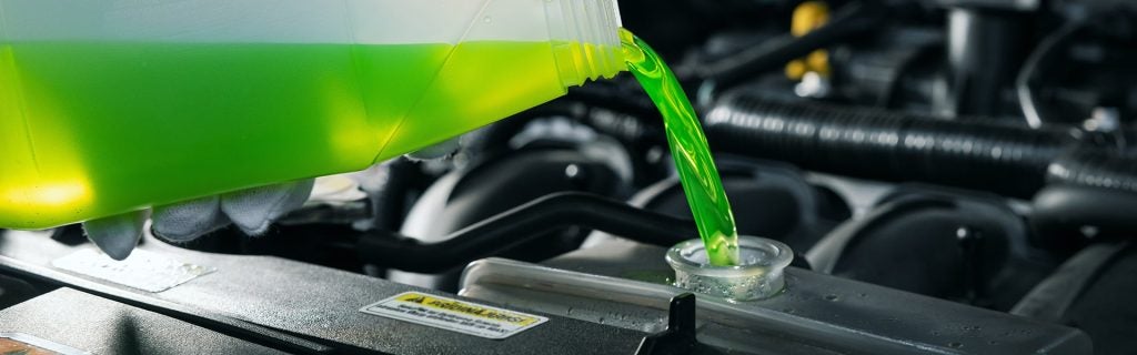 Bright green coolant pouring from a plastic bottle into a car's radiator, showing the engine compartment.