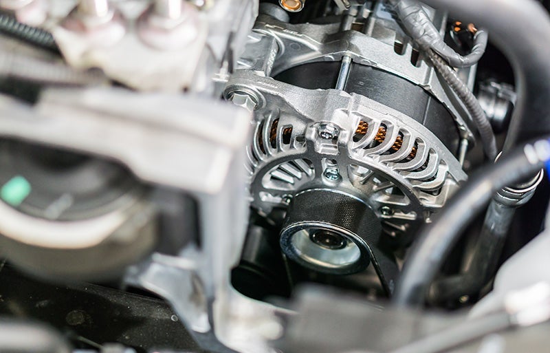 Close-up of a new car alternator with its pulley and drive belt, wires, and engine components.
