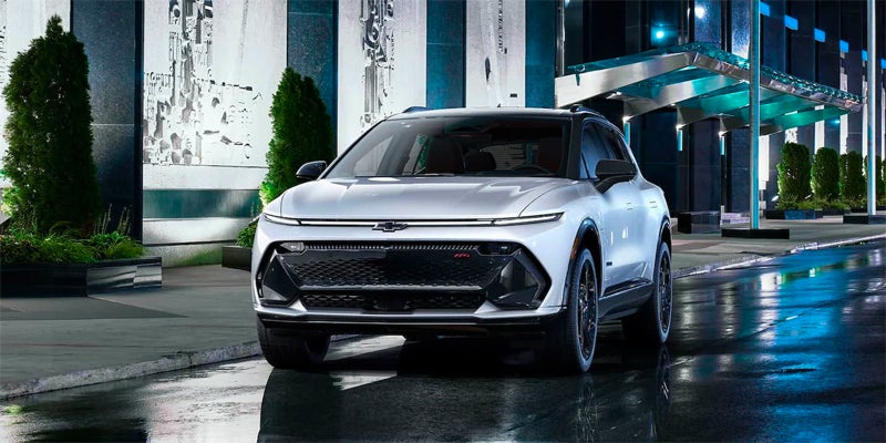Front view of a light gray Chevy Blazer EV parked at night, with reflections on the wet asphalt under blue lights.