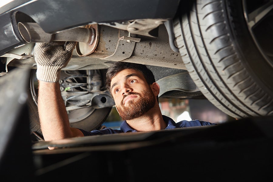 A gloved mechanic with a beard looks up at the underside of a car, adjusting its exhaust pipe.