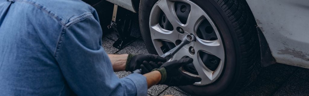 Someone in a denim jacket and gloves, kneeling to loosen lug nuts on a car tire with a wrench. A car jack is visible.